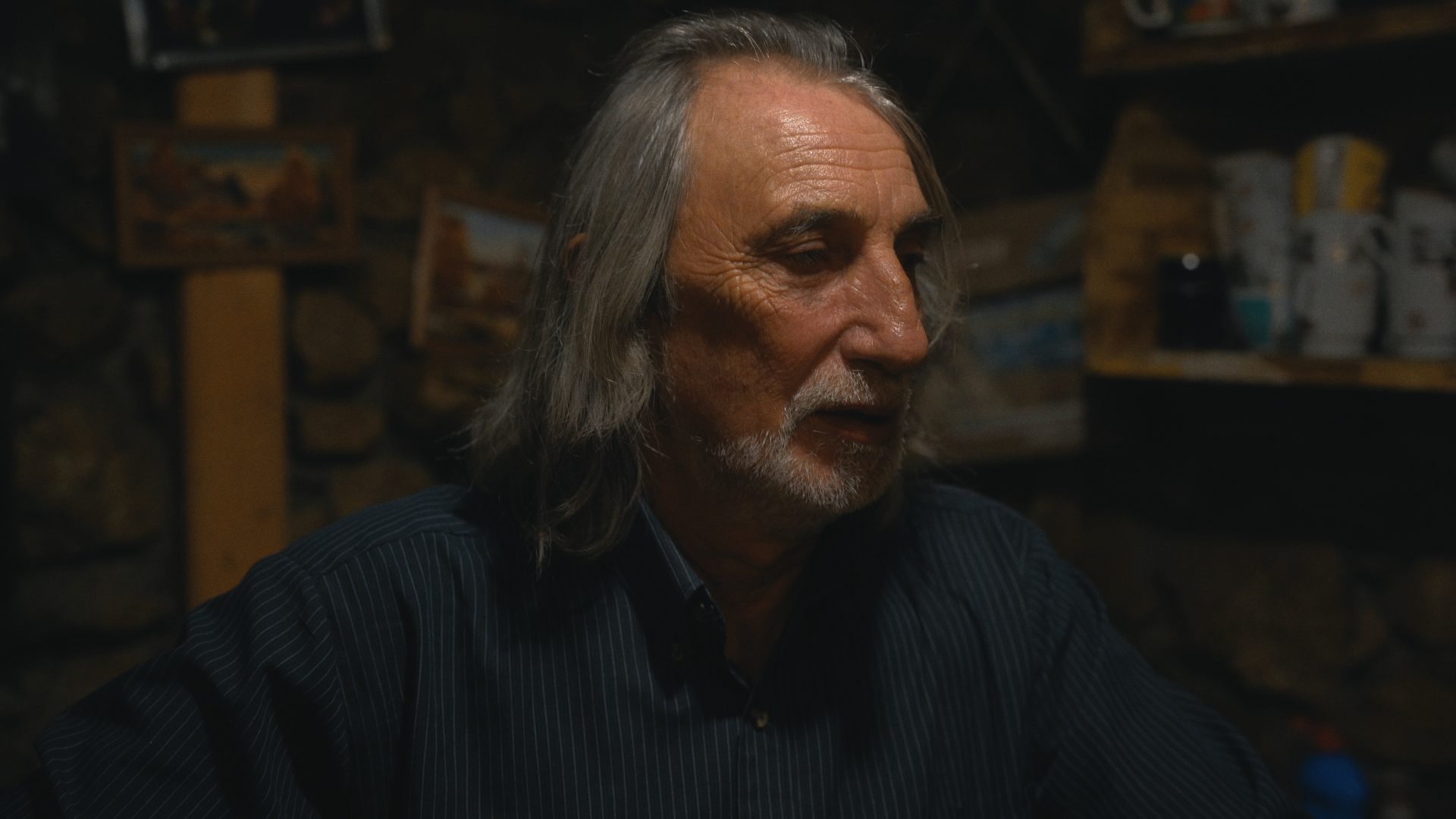 Älterer Mann mit grauem Haar und Bart sitzt in schwach beleuchtetem Raum mit Bildern im Hintergrund.