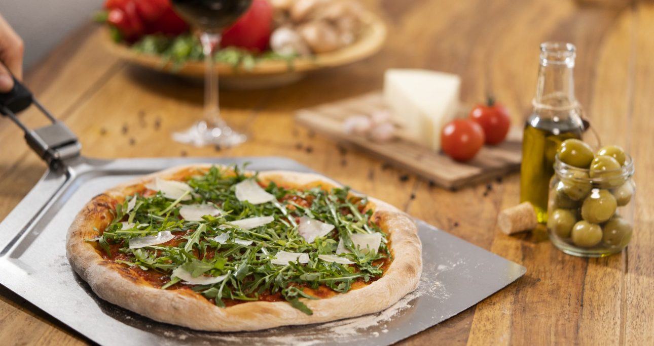Frische Pizza mit Rucola und Käse auf Platte, Tisch mit Rotwein, Oliven, Käse, Tomaten, Olivenöl.