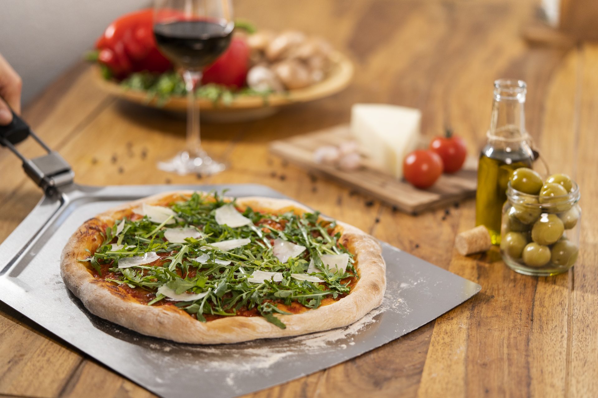 Frische Pizza mit Rucola und Käse auf Platte, Tisch mit Rotwein, Oliven, Käse, Tomaten, Olivenöl.