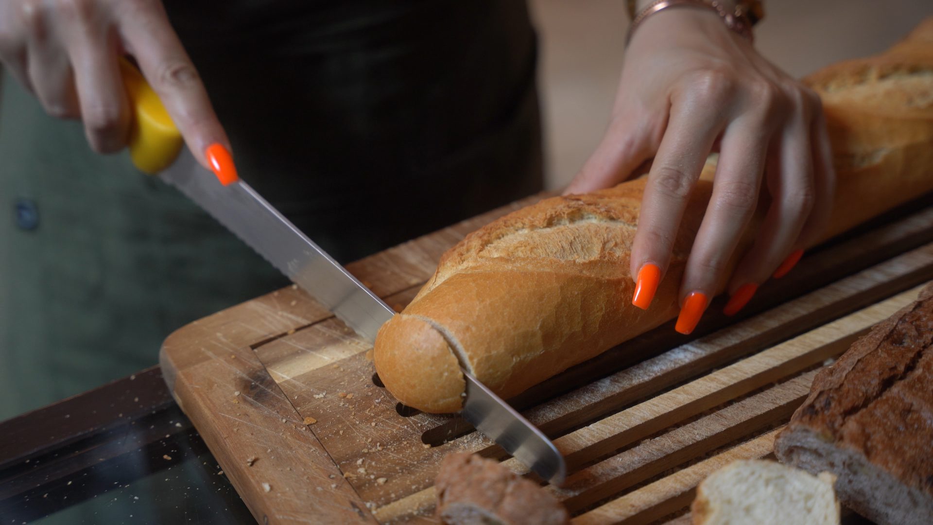 Person mit orangefarbenen Nägeln schneidet Baguette auf Holzbrett mit gezacktem Messer.