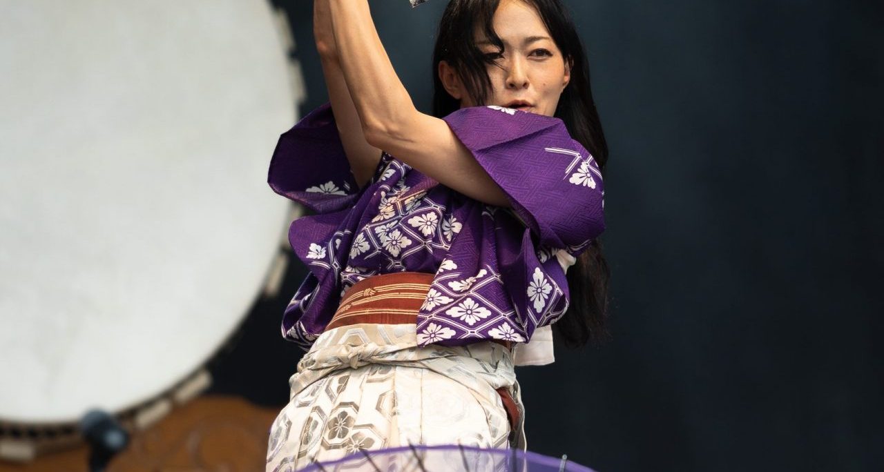 Eine Person in japanischer Kleidung tanzt mit Fächer, dekorativer Schirm im Vordergrund.