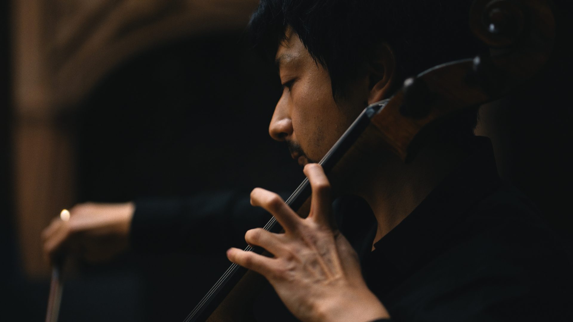 Eine Person spielt Cello, Fokus auf Griffbrett, Hintergrund dunkel beleuchtet.