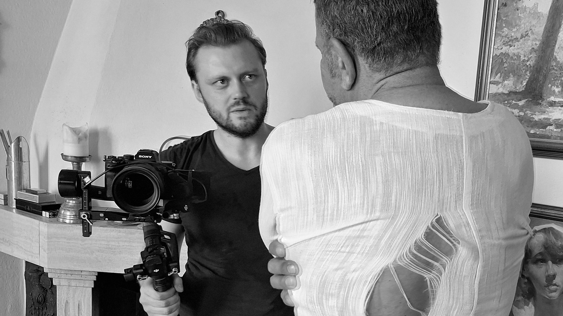 Zwei Personen im Gespräch, eine mit Kamera, weiße Kleidung, Kerze und Gemälde im Hintergrund.