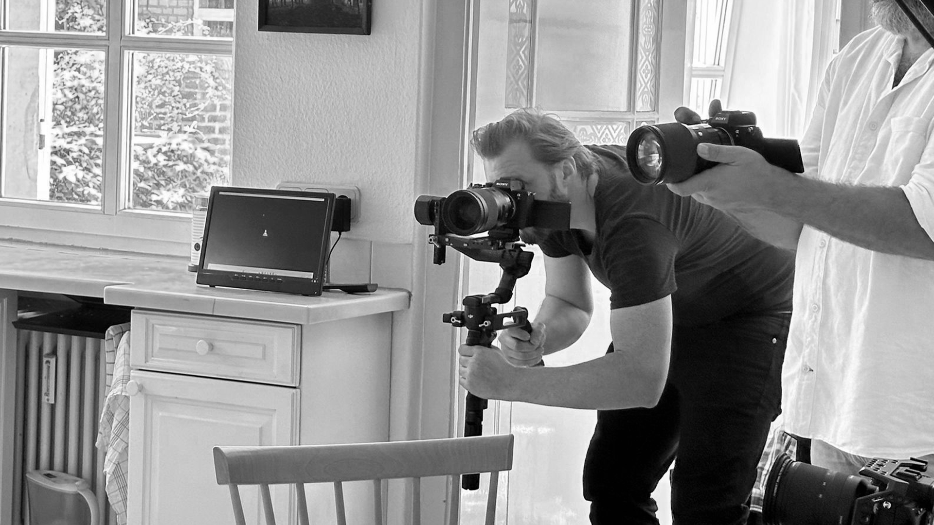 Zwei Personen bedienen Kameras in einem hellen Raum mit Fenstern und Monitor im Hintergrund.